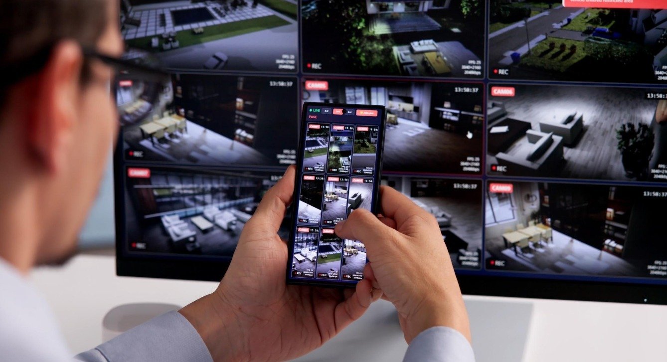 Authorised user reviewing camera feeds on a phone beside CCTV monitors